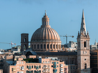 Fototapeta premium Valletta, Malta cityscape shows basilica dome and pro cathedral spire above limestone buildings, rooftop terraces, and cranes in clear daylight from an elevated vantage.