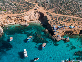 Aerial view shows Blue Lagoon on Comino Island, Malta. Turquoise water, honey colored limestone cliffs, beach umbrellas, tour boats, and dinghies in midday sun.