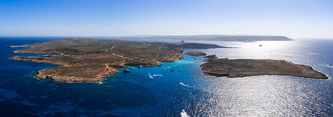 Aerial view shows Comino and Cominotto with the Blue Lagoon, boats on clear water, limestone terrain, and distant Gozo and Malta on the horizon in bright midday light.