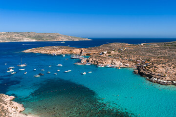 Aerial view of Blue Lagoon in Malta shows Comino and Cominotto, anchored boats, swimmers, and a narrow channel in bright midday light with vivid blue water