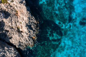 Aerial high angle shows Comino, Malta limestone ledges beside Blue Lagoon, turquoise to cobalt water, midday light, visible crevices, seagrass patches, and cave hint.