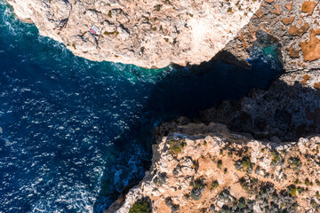 Aerial high angle shows Comino Island, Malta, with rugged limestone cliffs and a narrow Blue Lagoon inlet. Midday light creates crisp reflections and deep shadows over karst.