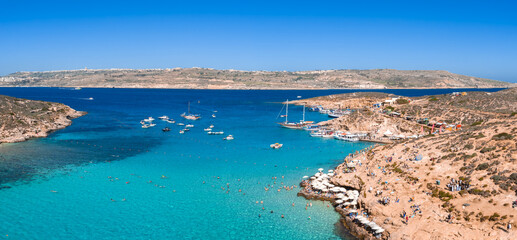 Aerial view of Blue Lagoon on Comino Island, Malta, with Gozo on the horizon. Boats anchor, swimmers and sunbathers gather by rocky coves under bright summer light.