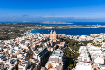 Aerial view shows Mellieha, Malta, with the red sandstone parish church above white buildings, Mellieha Bay, and Comino and Gozo on the horizon in bright midday light. © Aerial Film Studio