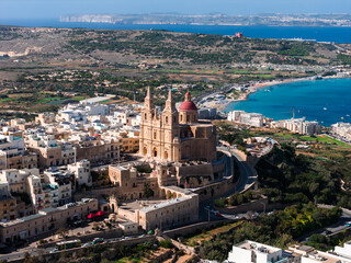 Naklejka premium Aerial view of the Parish Church of Mellieha with twin spires and red dome above limestone buildings, Mellieha, Malta. Mellieha Bay, Comino, and Gozo appear in clear daylight.