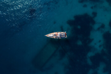 Directly above, a single sailboat rests on deep blue water off Sliema, Malta. Sun glints on the sea, reefs tint patches, and the hull casts a long diagonal shadow. © Aerial Film Studio