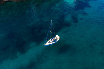 Aerial view shows a white sailboat anchored in clear teal to deep blue water off Sliema, Malta. Mast and rigging cast soft shadows over sand and darker patches. © Aerial Film Studio
