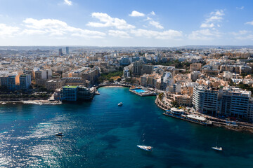 Fototapeta premium Aerial view of Sliema, Malta shows curving coastline, blue inlet, lido pool, boats, marinas, and waterfront apartments under midday light across the peninsula toward St. Julian's.