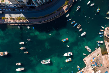 Aerial view of Sliema, Malta shows turquoise inlet with anchored boats, curving promenade, apartment blocks, roadway, and quay with slips in crisp midday light.