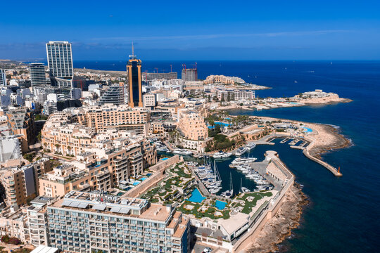 Aerial view of Sliema and St. Julian's, Malta, shows Portomaso Marina, the cylindrical Portomaso Business Tower, rocky peninsula, bays, pools, promenades, and beaches at midday.