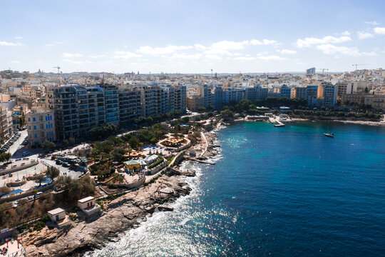 Aerial view of Sliema, Malta, in bright midday light. Rocky shoreline, modern mid rise blocks, promenade, terraced parks, and small boats curve along a calm bay.