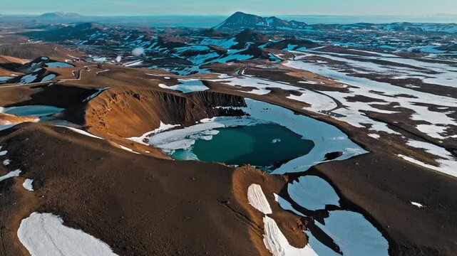 Drone footage of Krafla crater lake in North Iceland. Ice fragments float on calm blue water beside brown volcanic walls.