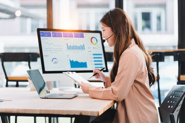 Young woman with headset analyzing business charts on monitor and laptop, taking notes with focused expression