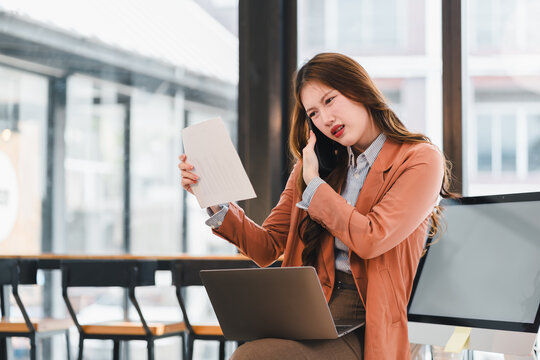 Young businesswoman on phone holds document and laptop, frustrated expression during office call and paperwork review