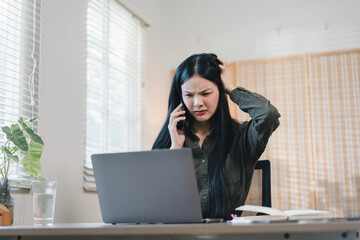 Frustrated woman on phone at home office with laptop and notebook, showing stress and concern while...