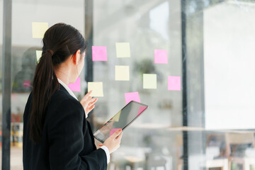 Young professional woman planning project with sticky notes on glass wall while holding tablet