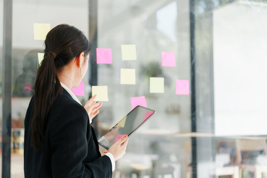 Young professional woman planning project with sticky notes on glass wall while holding tablet