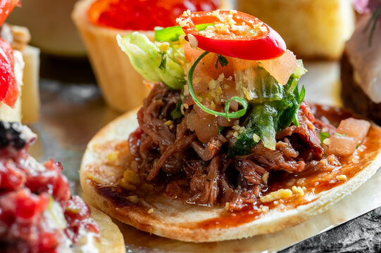 A closeup of a savory Mexican dish, possibly a chalupa or tostada, topped with slow-cooked shredded meat, fresh lettuce, red peppers, and a rich sauce on a toasted tortilla