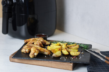 Air fryer with grilled chicken wings, potato wedges and spices on white marble countertop, closeup