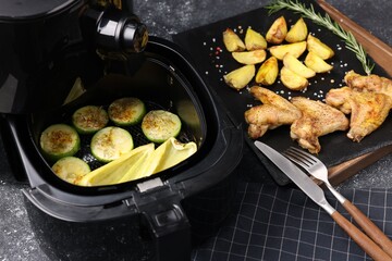 Air fryer with grilled vegetables, chicken wings and potato wedges on grey table, closeup