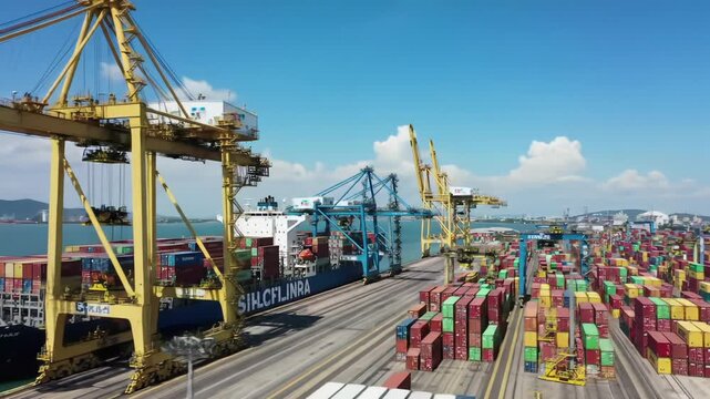 Sweeping aerial video of vast global container port at peak activity. Colossal blue ship docks as yellow/blue gantry cranes load colorful containers under clear sky, crisp mid-day sun. Concept of