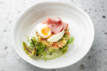 Gourmet dish with a soft-boiled egg, ham, peas, and mixed diced vegetables in a creamy sauce, garnished with fresh greens on a modern white plate