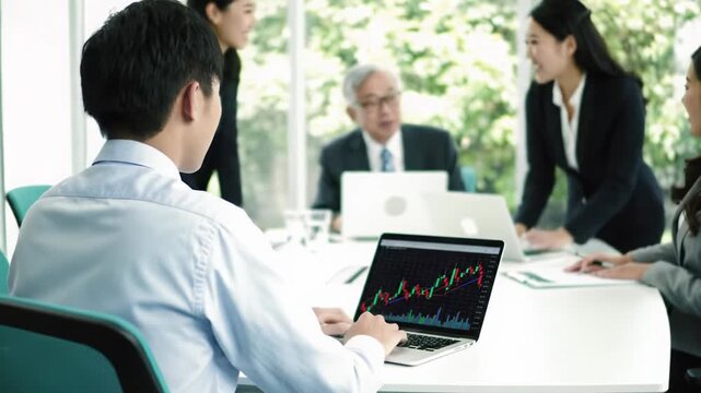 Diverse Asian professionals in suits collaborate at office table. Young man types financial graphs on laptop, blurred foliage. Intelligent collaboration and progress