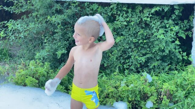 A happy young boy in bright yellow swim shorts is standing in white foam, smiling as he plays with a large bubble of foam on his head. He is enjoying the fun outdoors