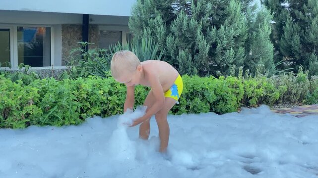 A young boy in yellow swim shorts is standing in a deep layer of white soap foam, holding a handful of it. He is playing an outdoor foam party surrounded by green bushes and a building.