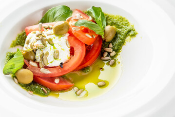 Fresh burrata cheese salad with sliced tomatoes, green olives, basil leaves, and sunflower seeds, drizzled with olive oil and green pesto on a white plate