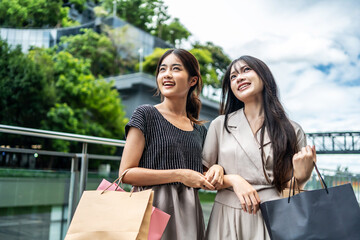 Portrait of a two happy young asian women friend holding shopping bags, standing together with happiness enjoy purchasing in front of the shopping mall center
