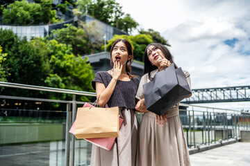 Portrait of a two happy young asian women friend holding shopping bags, standing together with happiness enjoy purchasing in front of the shopping mall center