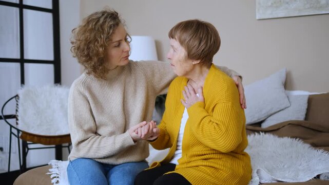 Young woman comforting her senior mother who is feeling unwell and experiencing discomfort. The granddaughter provides emotional support, holding her grandmother's hand in a caring way