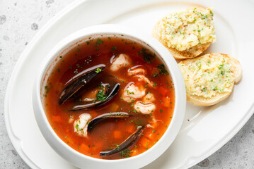 white bowl filled with a red seafood stew, including mussels and shrimp, garnished with fresh parsley. The bowl is on a white plate with two pieces of toasted bread topped with a savory spread