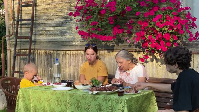 A family of three generations is sharing a lively outdoor summer meal of shashlik, flatbread, and fresh food. They are gathered around a table outside a rustic wooden house