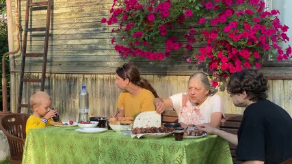 A family group of three generations is sharing an outdoor dinner featuring shashlik and flatbread. They are happily gathered around a table outside a rustic wooden dacha adorned with vibrant petunias