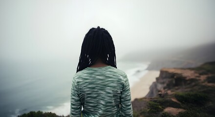 African American woman with dreadlocks standing on misty cliff edge looking at ocean view.