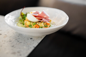 A dish of Olivier salad with ham and a half-boiled egg is served on a white plate on a modern countertop, captured in a restaurant setting
