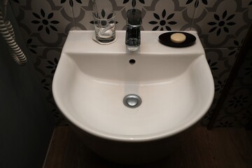Minimalist white ceramic bathroom sink with a bar of soap, glass, and water bottle placed on top, set against a patterned tile wall.
