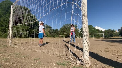 A young boy stands inside a football goal as his mother holds the ball, ready to play. They are enjoying a fun, casual game together on a dry field in a park on a sunny day.