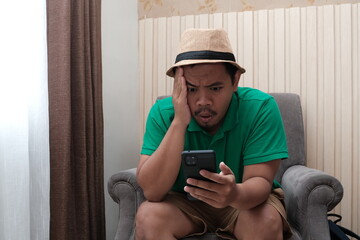Concerned man in a hat and green shirt sits on a chair, holding his head while looking at his...