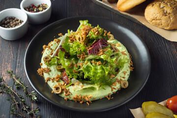 Fresh green salad with mix of lettuce, chard, olives and crispy topping served with green avocado or yogurt sauce in black plate on dark wooden table, healthy gourmet restaurant meal close-up