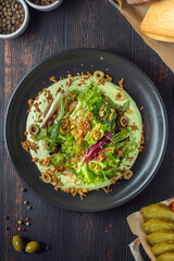 Fresh green salad with mix of lettuce, chard, olives and crispy topping served with green avocado or yogurt sauce in black plate on dark wooden table, healthy gourmet restaurant meal close-up