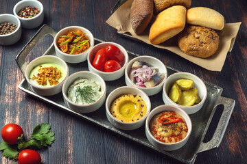 Assorted appetizers in small white bowls on metal tray, variety of dips, fish, vegetables and spreads served with fresh bread, meze sharing platter or tapas concept, top view on rustic table