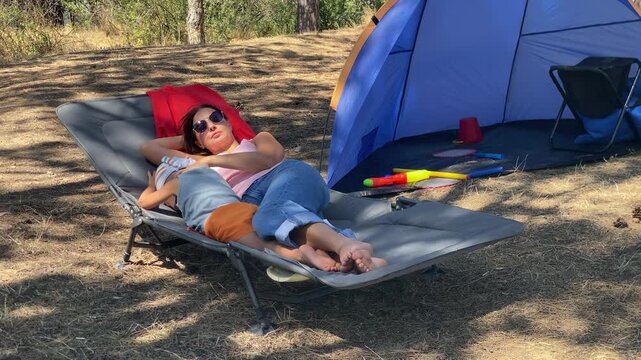A mother and her young son are lying closely on a camping cot, enjoying a moment of rest. They are at their campsite next to a blue tent in a sunny pine forest.