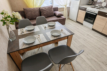 Cozy interior showcasing a dining area with a set table, surrounded by a sofa, kitchen appliances, and flowers. Warm lighting enhances the inviting ambiance