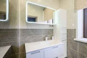 Modern bathroom featuring grey tiles, a white vanity with an integrated sink, a lighted mirror, and a tall storage cabinet