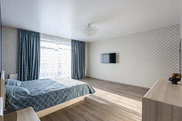Bright bedroom with blue bedding and curtains. A TV hangs on the textured wall with sunlight flooding through sheer window coverings