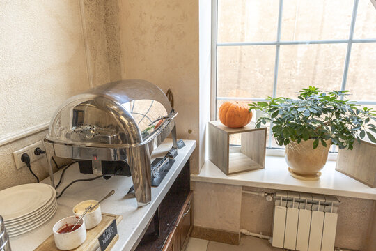 Hotel breakfast buffet with chrome food warmer, plates, small bowls of sauce, and a lush plant and a pumpkin on a sunlit windowsill, creating a cozy scene.