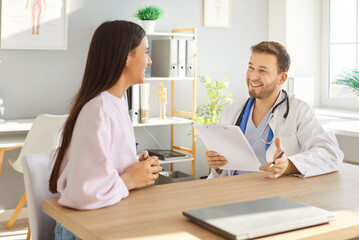 Fototapeta premium Portrait of a young woman visiting friendly male doctor sitting at the desk in clinic during medical examination. Female patient talking with physician in office. Health care and medicine concept.